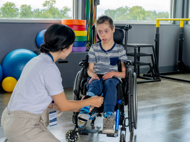 A physiotherapist puts on a young boys orthopedic braces as they prepare to being a therapy session together.  The boy is seated in his wheelchair and is watching the therapist closely.